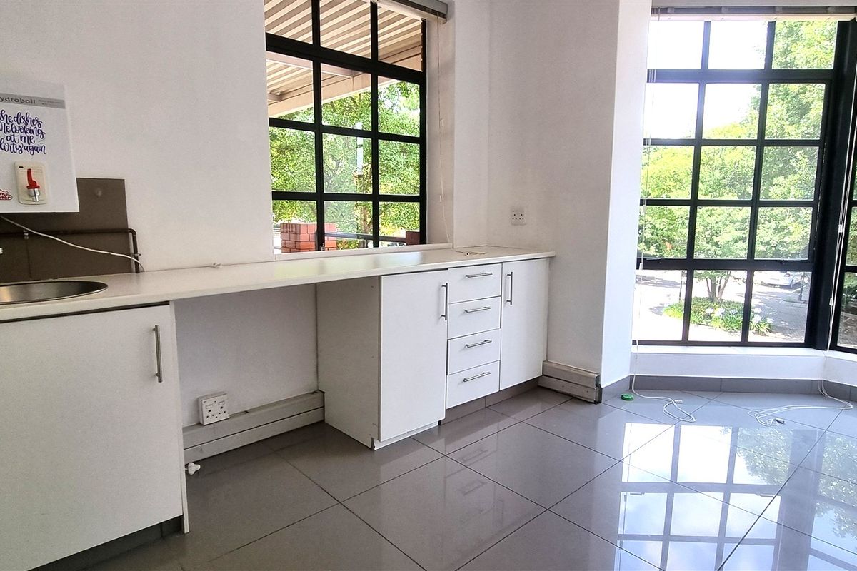 A bright office kitchenette area featuring white cabinetry, a stainless steel sink, a wall-mounted water boiler, and large windows with black frames.