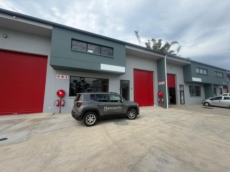 Modern industrial unit exterior featuring prominent red roller shutter doors, fire safety equipment, and a clean concrete driveway approach.