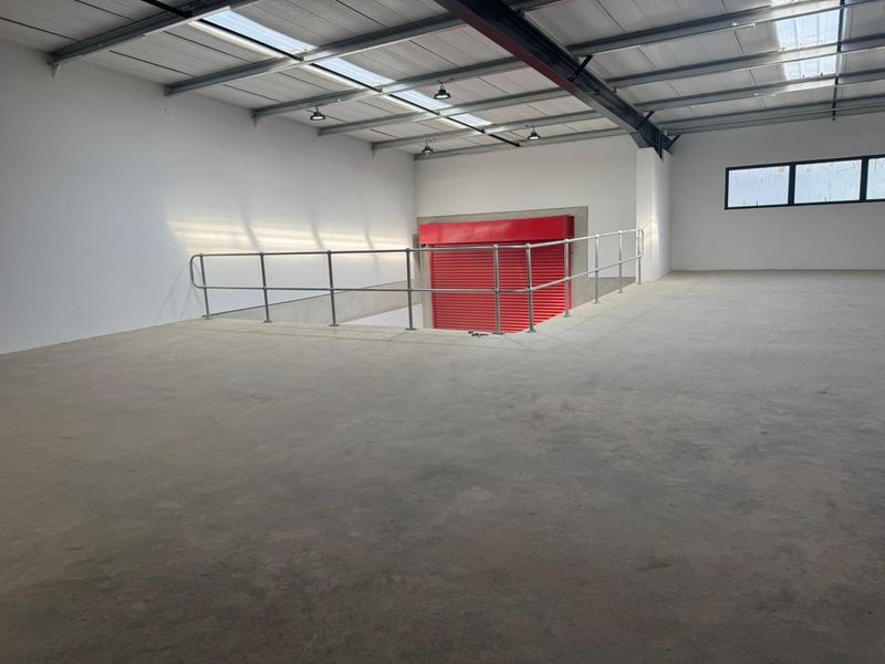 Spacious industrial mezzanine area featuring concrete flooring, exposed steel roof trusses, skylights, and a secure red metal roller shutter door.