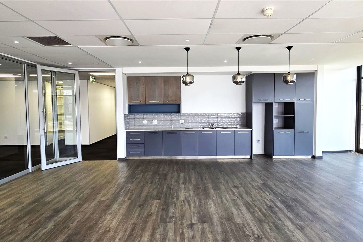 Modern office kitchenette featuring dark grey cabinetry, wood-tone upper cabinets, subway tile backsplash, and integrated sink, set within an open-plan workspace.
