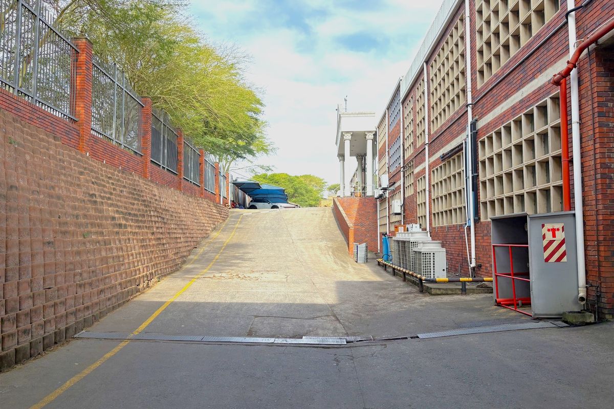 Industrial exterior view featuring a concrete access ramp, brick retaining wall with metal fencing, and multi-level brick building with grid-patterned window screens.