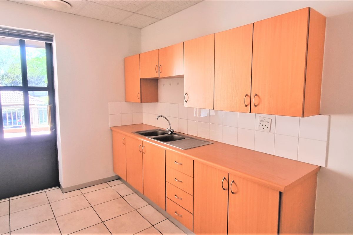 A functional office kitchenette featuring light wood-finish cabinetry, a stainless steel double sink, tiled backsplash, and direct access to an exterior door.