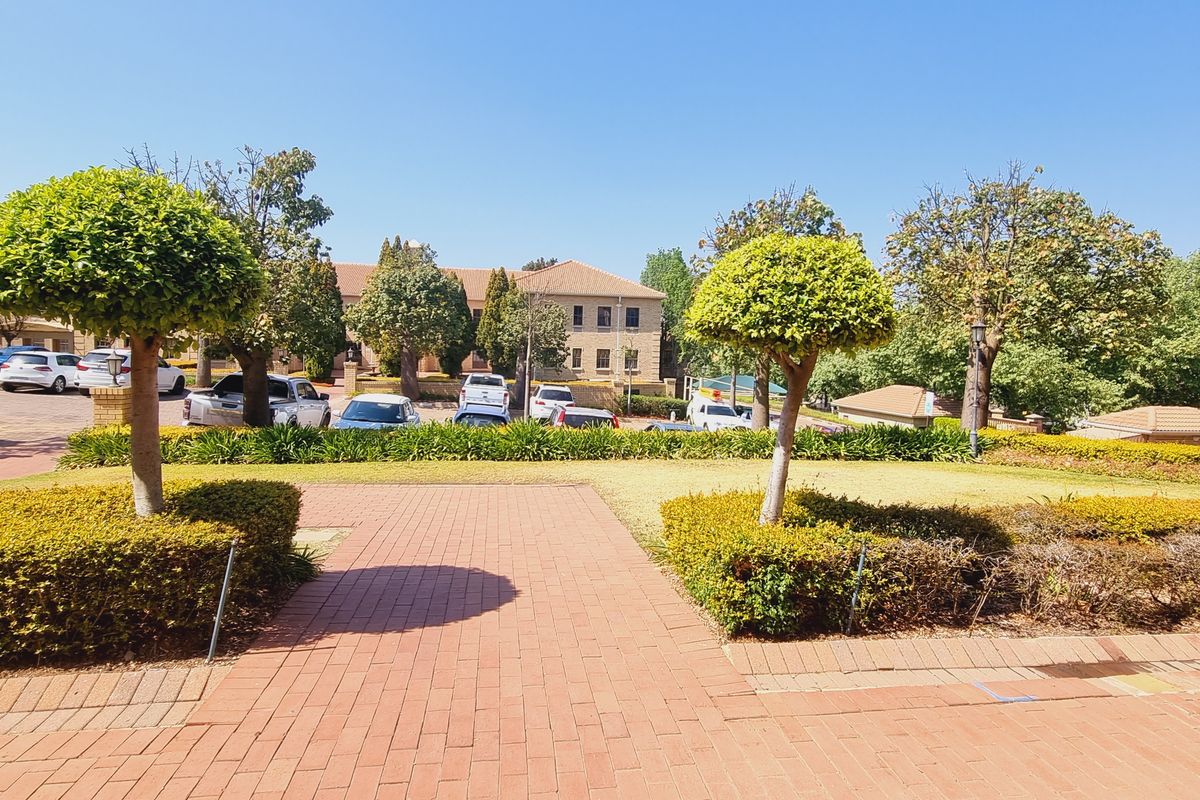 A well-maintained office park exterior featuring paved walkways, manicured hedges, and mature trees leading toward the main office building and parking area.