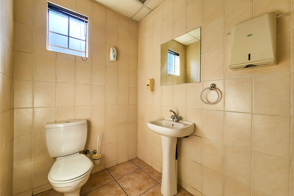 A functional office restroom featuring a pedestal sink, toilet, wall-mounted paper towel dispenser, and full-height wall tiling.