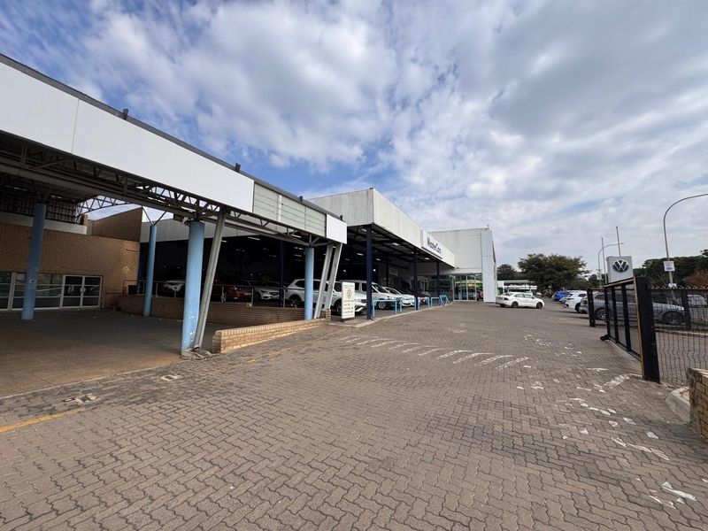 Front exterior view of a retail car dealership featuring a large paved forecourt, covered display areas, and steel structural canopy.