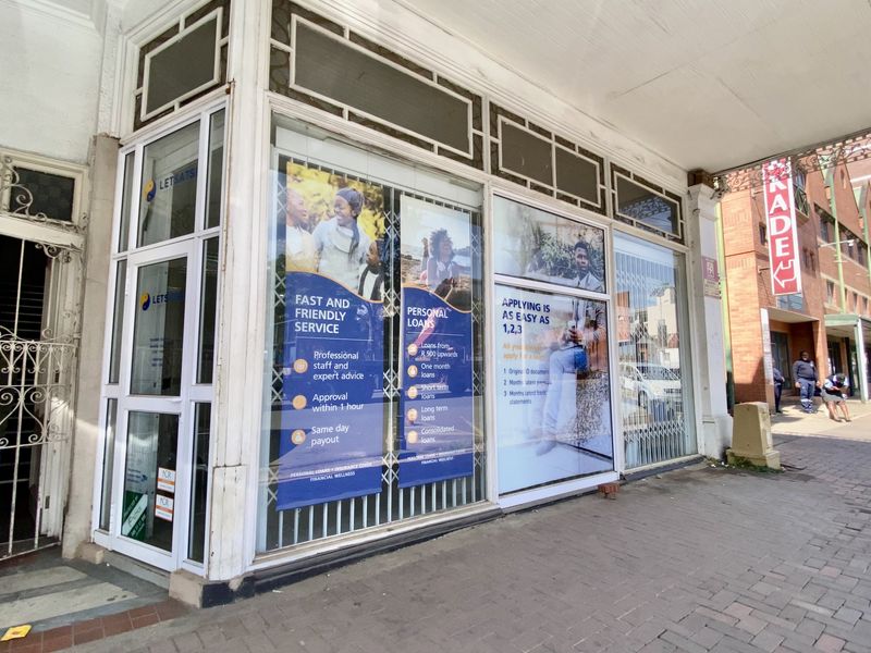 Commercial storefront featuring large display windows, a covered walkway, and metal security gate access in a high-traffic street setting.