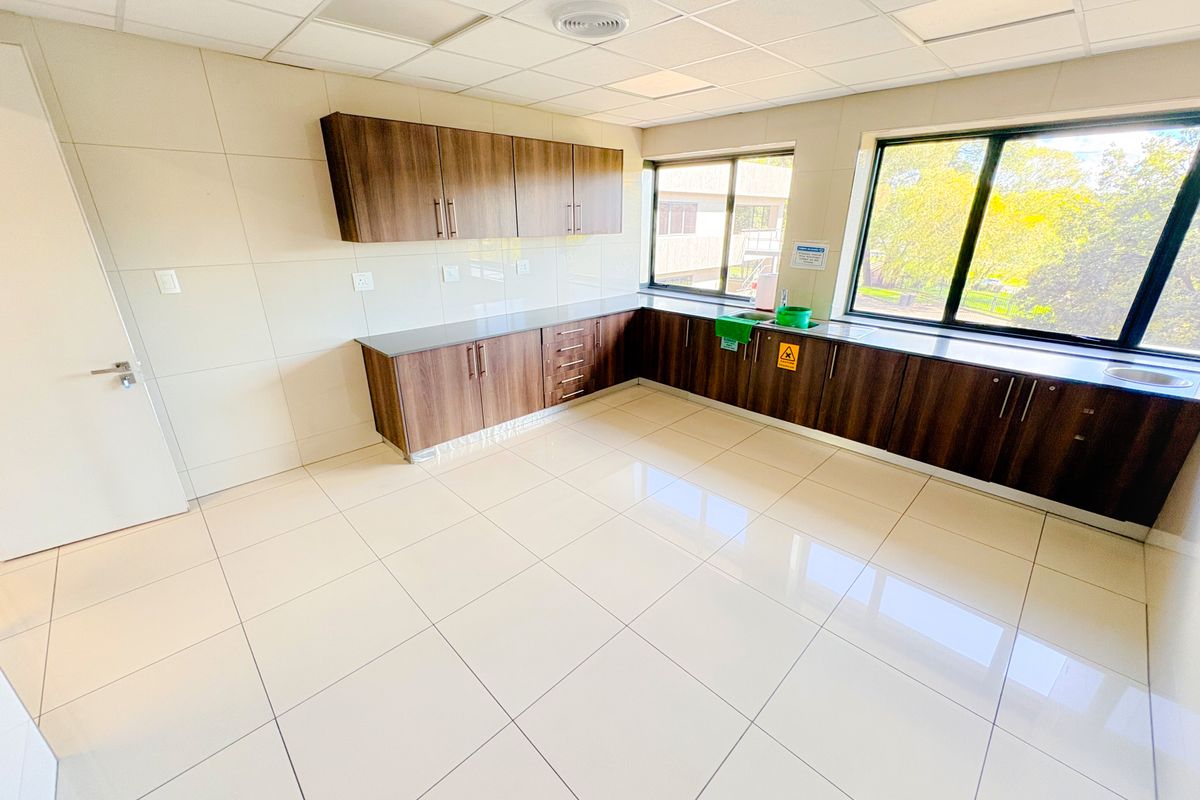 A clean, modern office kitchenette featuring dark wood cabinetry, stone countertops, tiled flooring, and large windows providing ample natural light.