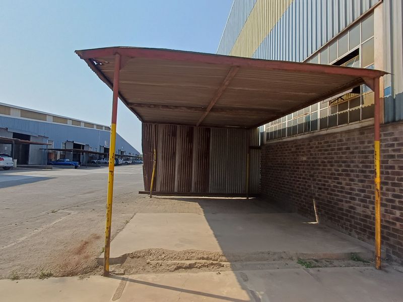 A weathered outdoor covered structure with a corrugated metal roof and steel supports, situated adjacent to a brick industrial building.
