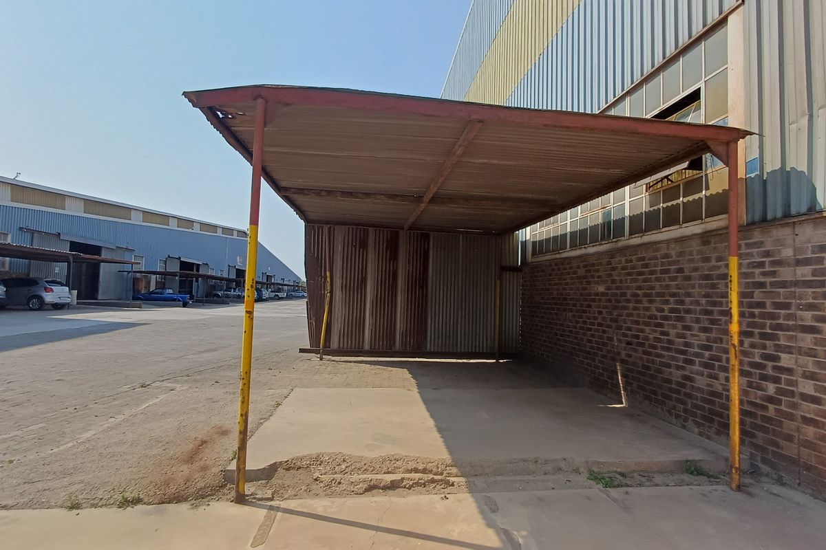 A weathered outdoor covered structure with a corrugated metal roof and steel supports, situated adjacent to a brick industrial building.