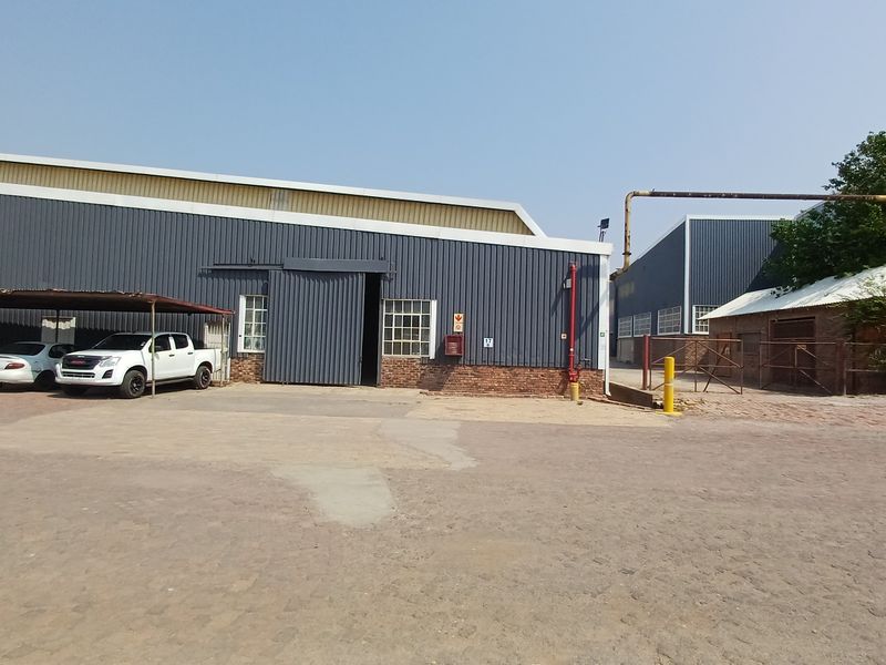 Industrial warehouse facility featuring corrugated metal cladding, a large sliding entrance door, brick base, and an attached steel carport structure.