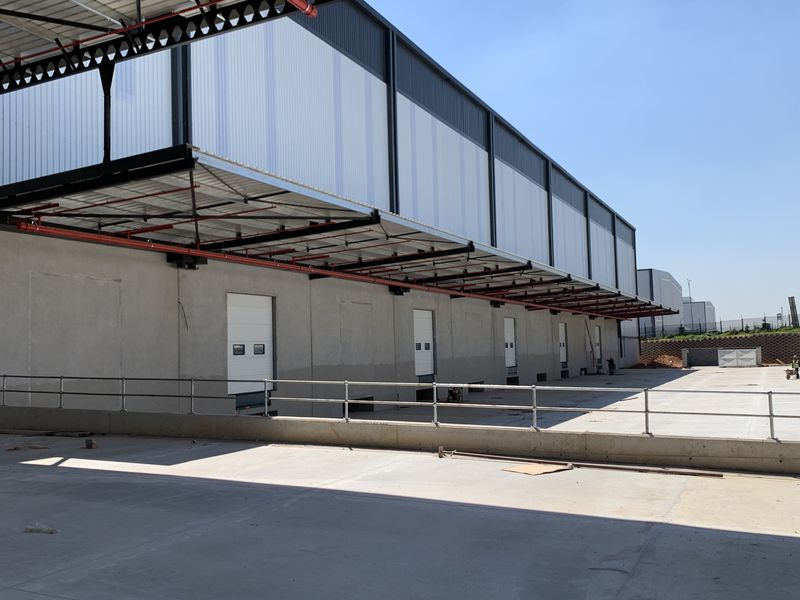Industrial warehouse exterior featuring a concrete loading yard, multiple roller shutter doors, and a large protective steel canopy.