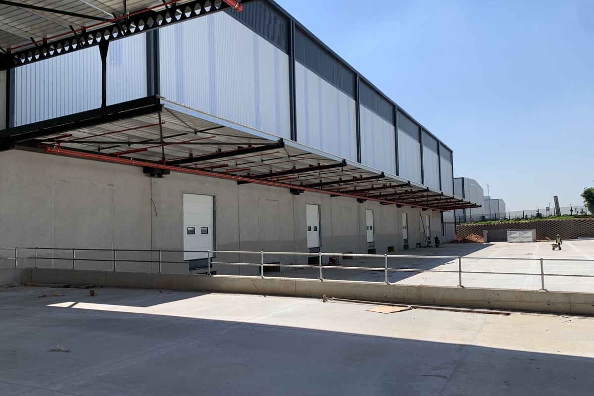Industrial warehouse exterior featuring a concrete loading yard, multiple roller shutter doors, and a large protective steel canopy.