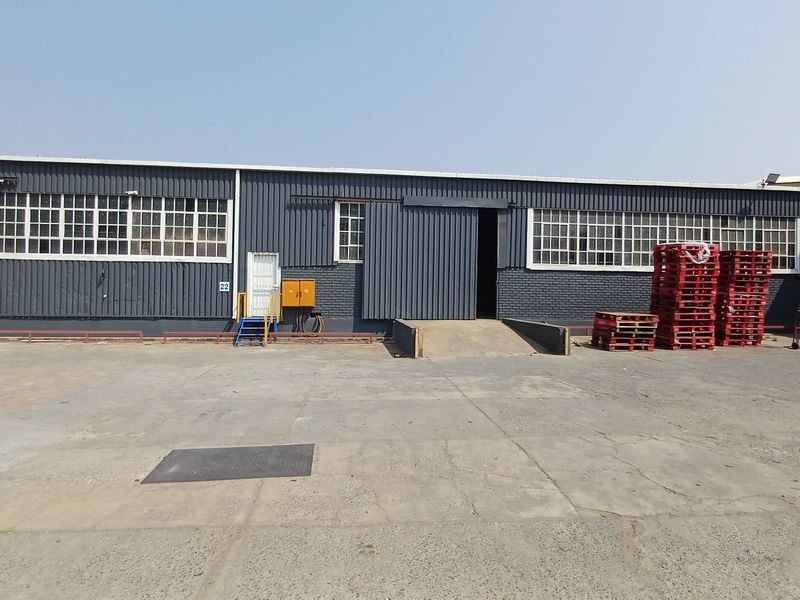 Industrial warehouse exterior featuring a large concrete yard, loading bay ramp, sliding access door, and extensive steel-framed window glazing.