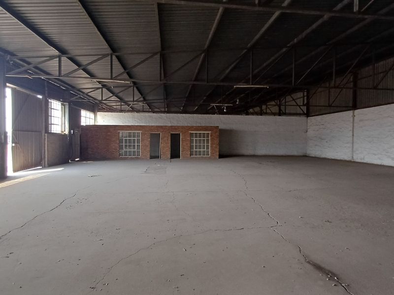 Spacious industrial warehouse featuring exposed steel roof trusses, concrete flooring with visible cracking, and a central brick partition wall with integrated windows.