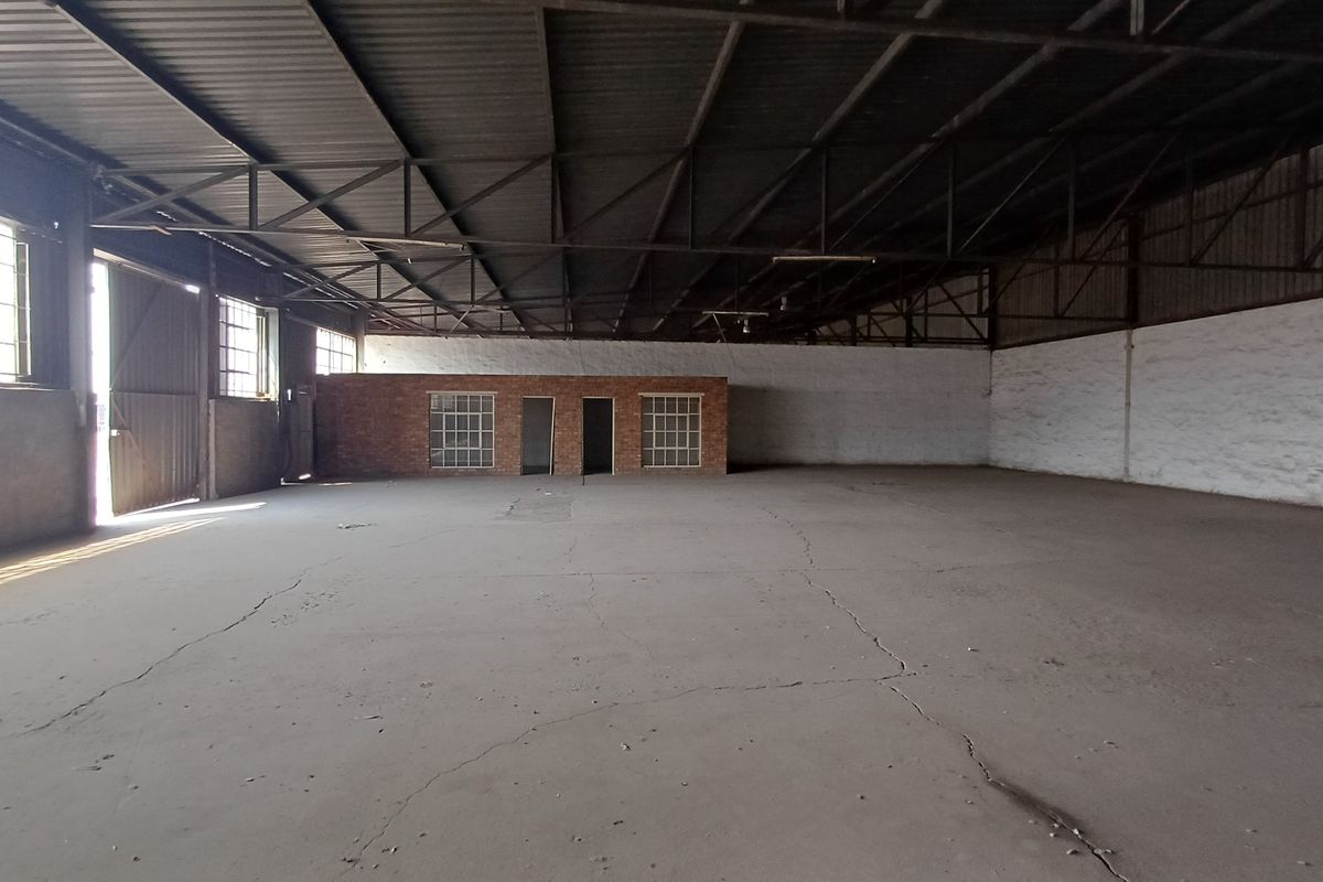 Spacious industrial warehouse featuring exposed steel roof trusses, concrete flooring with visible cracking, and a central brick partition wall with integrated windows.