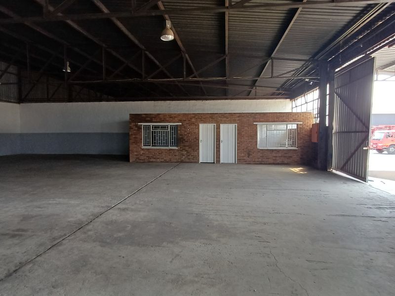 Spacious industrial warehouse featuring high ceilings with exposed steel trusses, concrete flooring, and a central brick office partition with secure access.