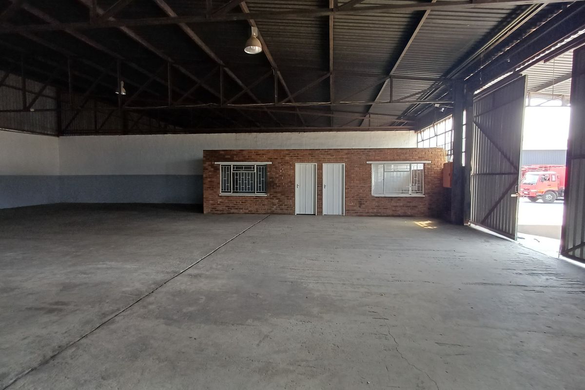 Spacious industrial warehouse featuring high ceilings with exposed steel trusses, concrete flooring, and a central brick office partition with secure access.
