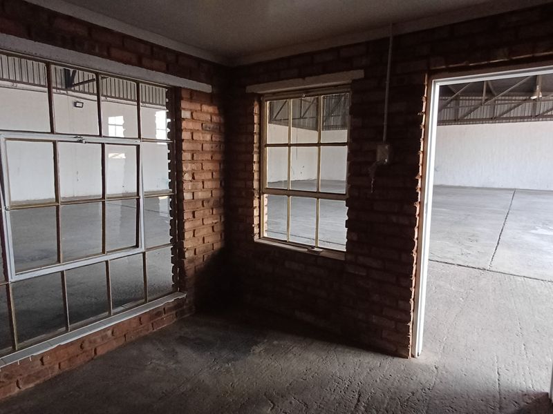 An empty industrial office space featuring exposed brick walls, concrete flooring, and steel-framed windows overlooking the main warehouse area.