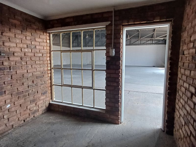 An empty office space featuring exposed brick walls, a large steel-framed window, and concrete flooring, providing a raw industrial aesthetic.