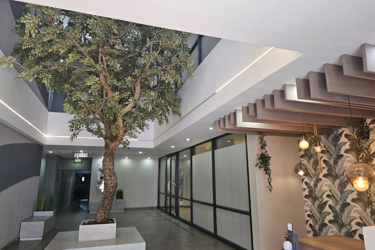 Modern office building entrance lobby featuring high ceilings, glass partitions, tiled flooring, and decorative timber ceiling accents with a central feature planter.