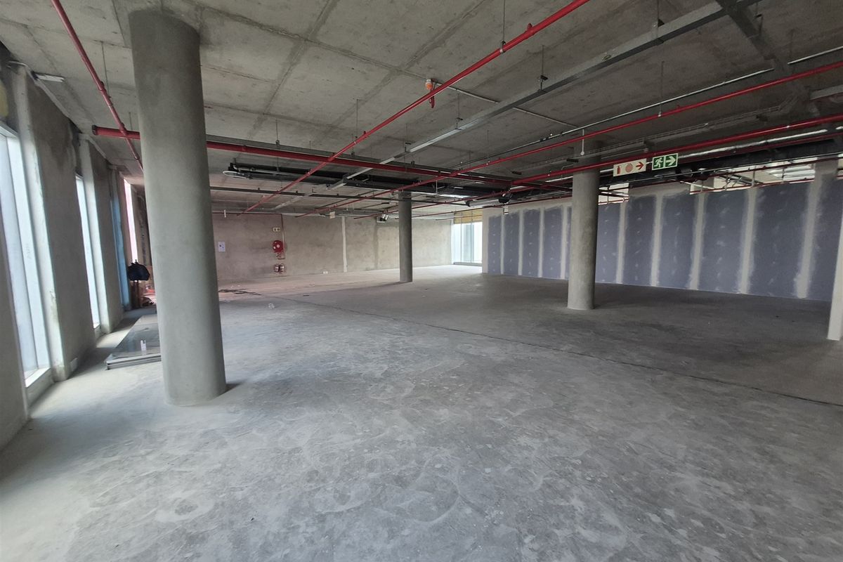 An expansive, open-plan office space in shell condition, featuring exposed concrete ceilings, structural columns, and large windows providing ample natural light.