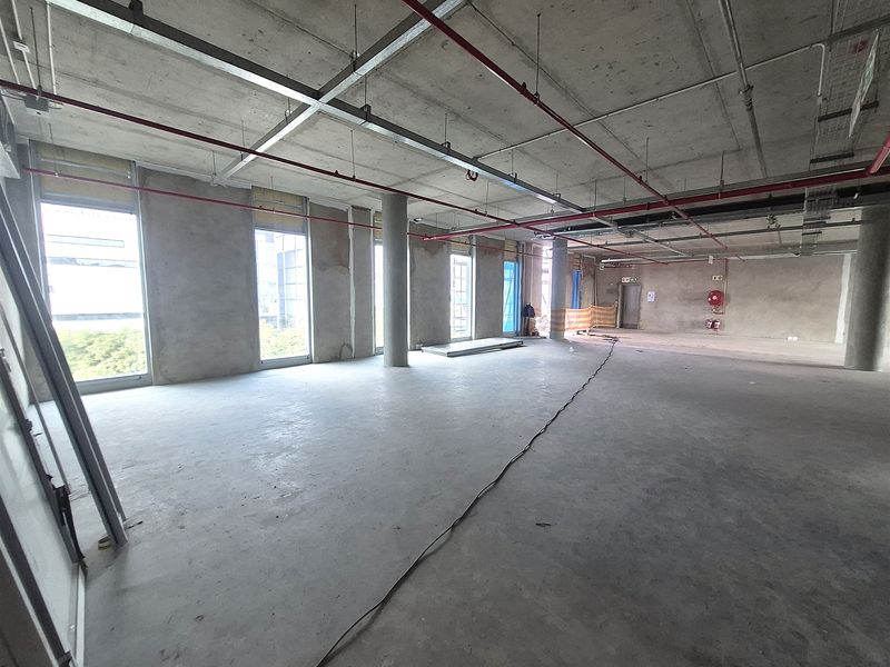 An expansive, unfinished open-plan office space featuring exposed concrete ceilings, structural columns, and large windows providing ample natural light.