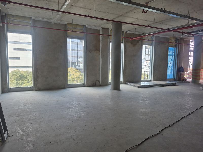 An unfinished open-plan office space featuring exposed concrete ceilings, structural columns, fire sprinkler systems, and large windows providing ample natural light.