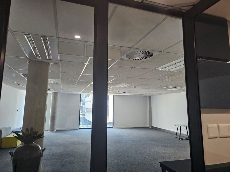 A spacious, open-plan office area featuring a suspended ceiling, recessed lighting, concrete support columns, and neutral carpet flooring, viewed through a glass partition.