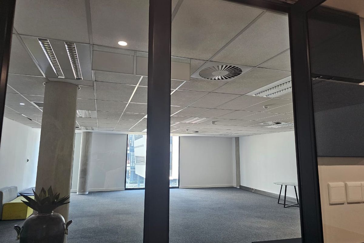A spacious, open-plan office area featuring a suspended ceiling, recessed lighting, concrete support columns, and neutral carpet flooring, viewed through a glass partition.
