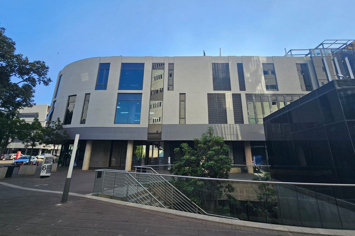 Modern office building exterior featuring a curved facade, large glass windows, louvred shutters, and a paved pedestrian entrance area.