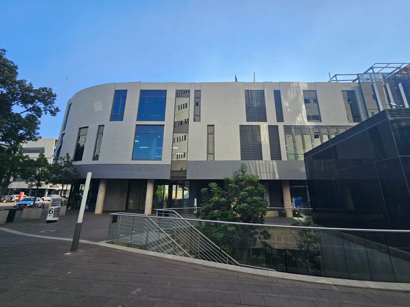 Modern office building exterior featuring a curved facade, large glass windows, louvred shutters, and a paved pedestrian entrance area.