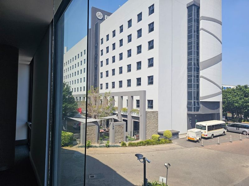 An elevated view looking out from an office window towards a modern multi-story commercial office complex and paved parking area.