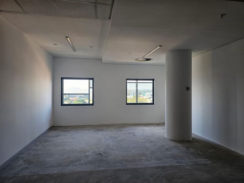An empty, open-plan office space featuring a structural column, suspended ceiling, and large windows with city views, awaiting final flooring installation.