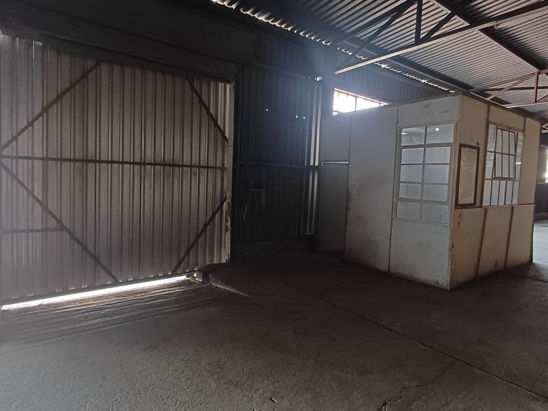 Spacious industrial warehouse area featuring high ceilings with exposed steel trusses, a large sliding metal door, and a partitioned internal office structure.