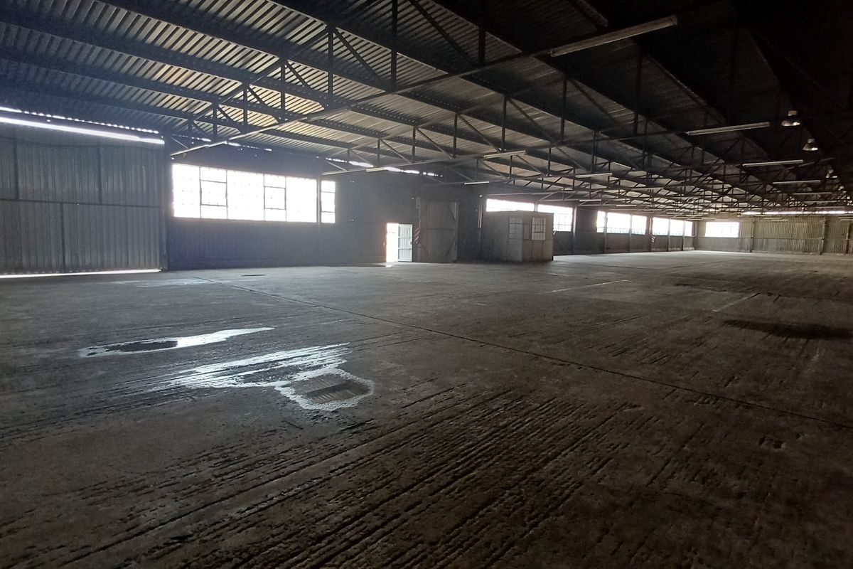 A large, open-plan industrial warehouse space featuring exposed steel roof trusses, concrete flooring, and high-level windows for natural light.