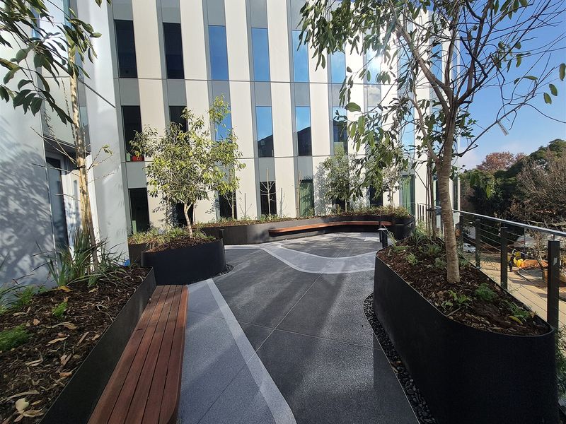 A modern outdoor terrace area featuring built-in timber bench seating, raised metal planters, and paved flooring, set against a contemporary office building facade.