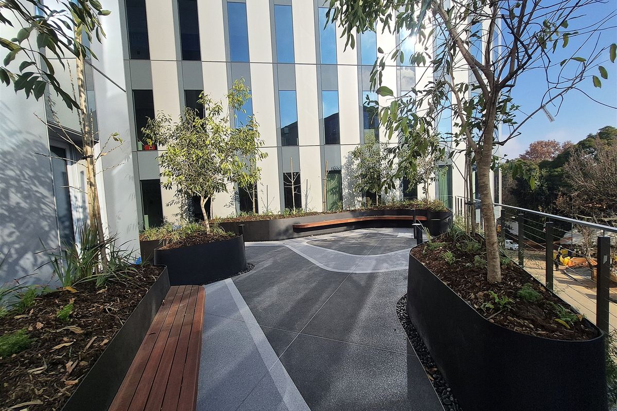 A modern outdoor terrace area featuring built-in timber bench seating, raised metal planters, and paved flooring, set against a contemporary office building facade.