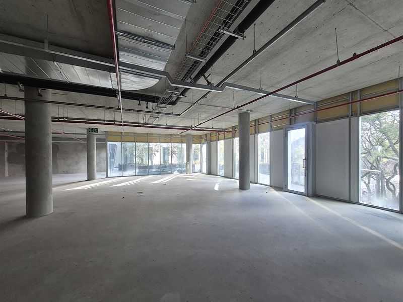 An open-plan office space in shell condition featuring exposed industrial ceilings, concrete columns, and expansive floor-to-ceiling windows providing natural light.