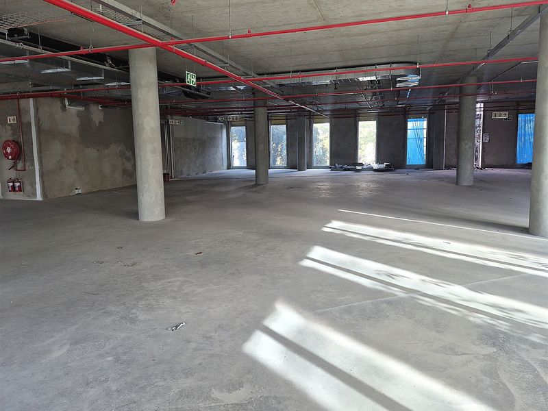 A large, open-plan office space in shell condition, featuring exposed concrete ceilings, structural columns, and visible fire safety infrastructure.