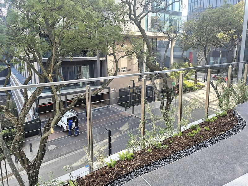 Elevated view overlooking the street entrance and surrounding office complex, featuring a modern stainless steel and wire cable balustrade with landscaped garden borders.