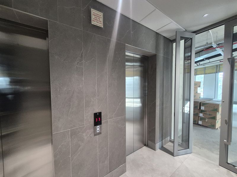 Modern office elevator lobby featuring sleek stainless steel lift doors and contemporary stone-look wall tiling, leading into an unfinished open-plan office space.
