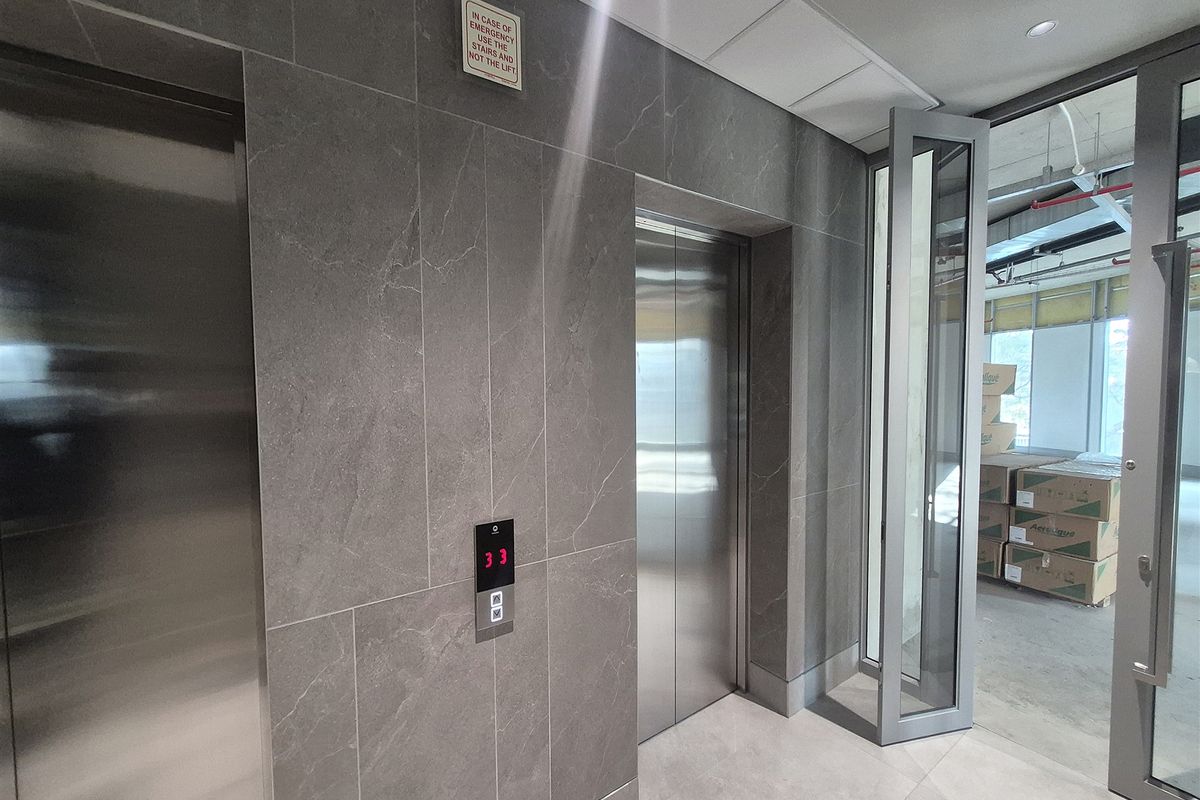 Modern office elevator lobby featuring sleek stainless steel lift doors and contemporary stone-look wall tiling, leading into an unfinished open-plan office space.