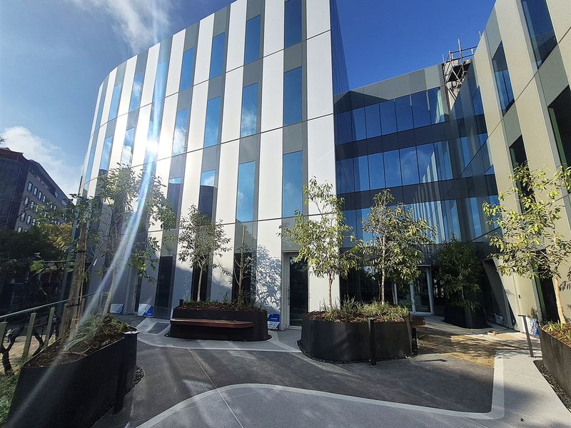 Modern office building exterior featuring a contemporary glass and metal facade with a landscaped paved courtyard and integrated planters.