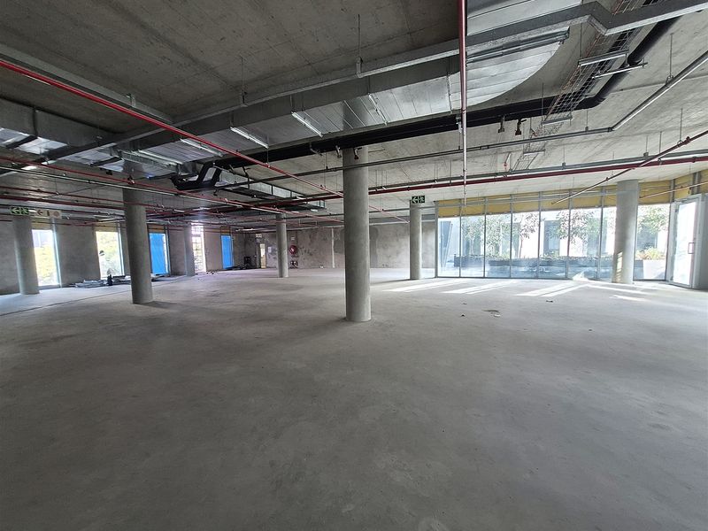 A large, open-plan office space in shell condition, featuring exposed concrete ceilings, industrial ducting, and expansive floor-to-ceiling windows providing natural light.