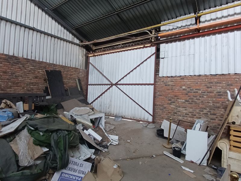 Industrial utility space featuring exposed brick walls, corrugated metal roofing, and a large sliding access door, currently requiring significant clearing and maintenance.