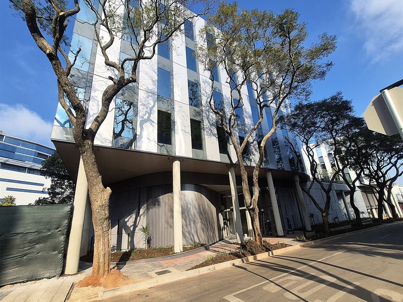 Modern multi-story office building featuring a glass curtain wall facade, structural support columns, and a paved entrance area.