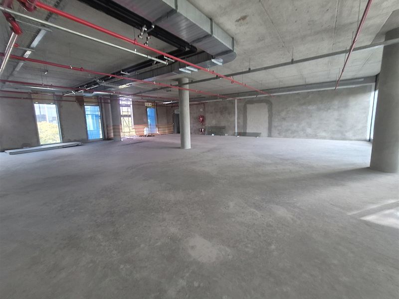 A large, open-plan office space in shell condition, featuring exposed concrete ceilings, industrial ducting, fire sprinklers, and expansive windows providing natural light.