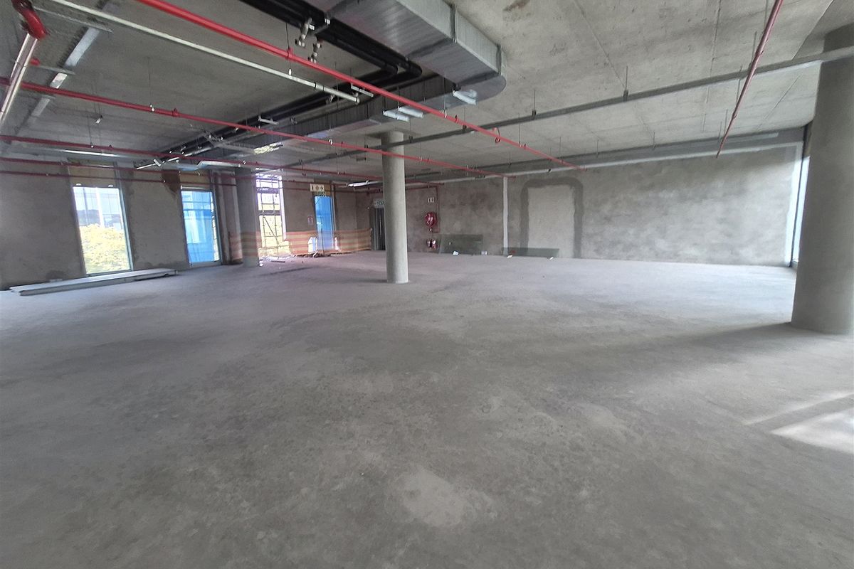 A large, open-plan office space in shell condition, featuring exposed concrete ceilings, industrial ducting, fire sprinklers, and expansive windows providing natural light.