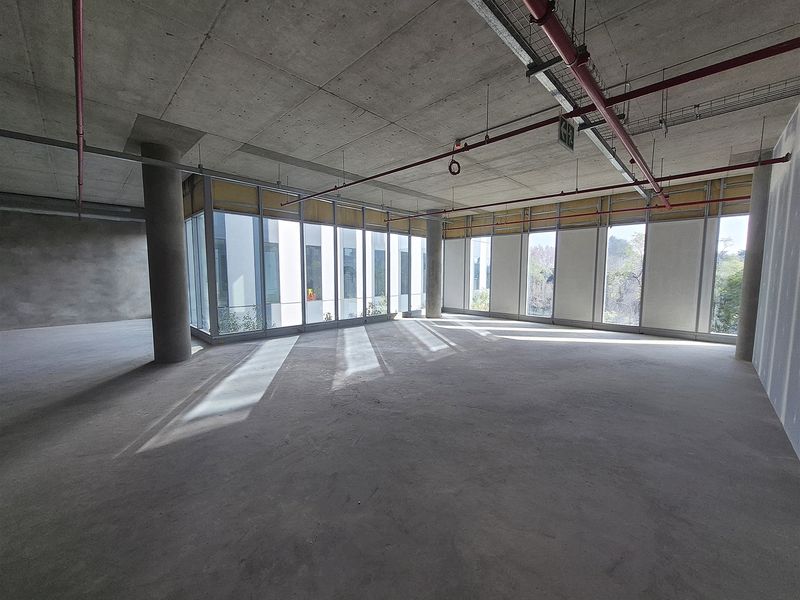 An expansive, open-plan office space in shell condition, featuring exposed concrete ceilings, industrial fire sprinkler systems, and large windows providing ample natural light.