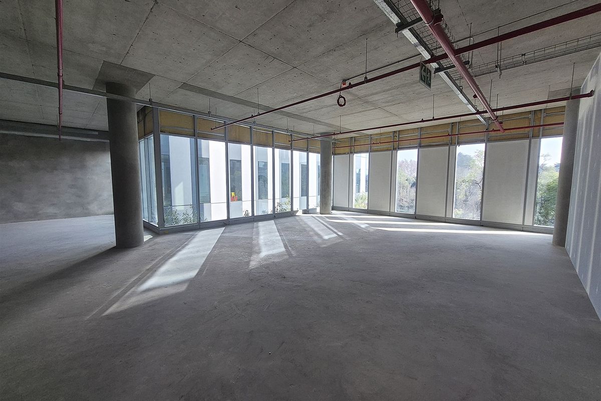 An expansive, open-plan office space in shell condition, featuring exposed concrete ceilings, industrial fire sprinkler systems, and large windows providing ample natural light.